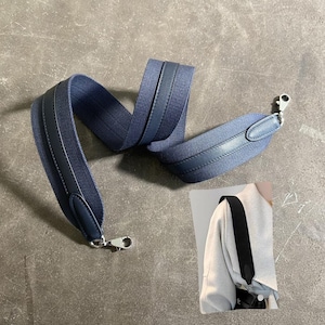 Puede incluir: Una correa de bolso de tela tejida azul con ribetes de cuero liso en los bordes y cierres de mosquetón de metal plateado en ambos extremos. Un recuadro muestra la correa unida a un bolso, demostrando su anchura y colocación sobre el hombro.