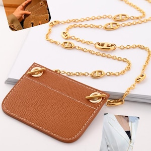 May include: A tan leather card holder with a textured surface and white stitching, featuring two gold-tone metal toggle closures at the top corners. A long gold-tone chain with oval link accents is attached to the toggles for carrying.