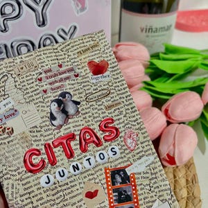 Puede incluir: Un álbum de recortes con las palabras "CITAS JUNTOS" en letras rojas, con pegatinas de pingüinos, fotos de tiras de película y la fecha 23-10-2025. Una botella de vino Viñamar y tulipanes rosas en el fondo.