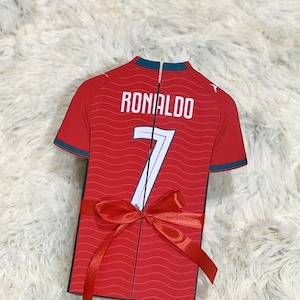 Puede incluir: Una caja de regalo con forma de camiseta roja que presenta un patrón de líneas onduladas, el nombre RONALDO y el número 7 en blanco. La caja tiene bordes verde oscuro en el cuello y las mangas, y está atada con un lazo rojo brillante al frente.