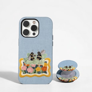 Puede incluir: Una funda de smartphone azul claro y un soporte circular para teléfono a juego. Ambos artículos presentan una ilustración de dos abejas sentadas en un sofá verde sobre una alfombra con flores rosas, amarillas y naranjas. La funda tiene una abertura.