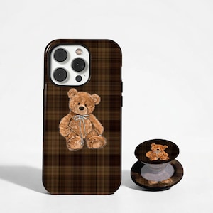 May include: A brown plaid phone case and matching phone grip featuring a teddy bear illustration. The phone case is designed to fit a smartphone and the grip is round with a collapsible stand.