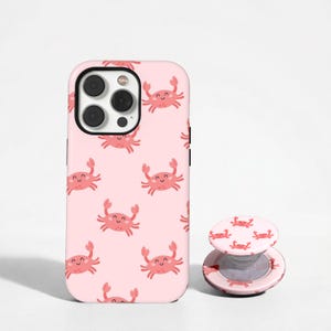 May include: Pink phone case and matching pop socket featuring a repeating pattern of smiling cartoon crabs. The phone case is on a white background. The pop socket is round and has a collapsible design.