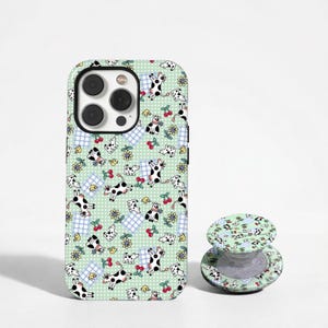 May include: A phone case and matching phone grip with a cow pattern on a pale green backdrop. The design features cartoon cows, cherries, and flowers. The phone case is black and white.
