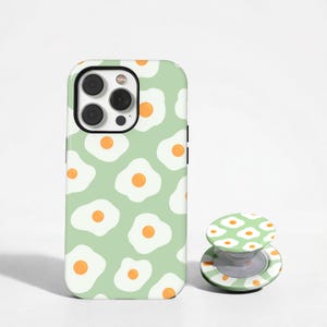 May include: A mint green phone case and matching phone grip featuring a repeating pattern of fried eggs with orange yolks. The phone case has a black camera lens border. The phone grip is round with a white and green striped base.