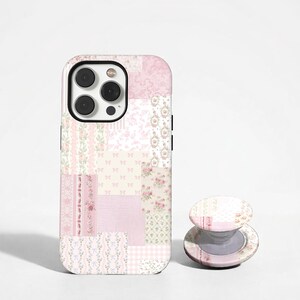 Pode incluir: Uma capa de telefone rosa em patchwork com uma pega pop-up a condizer. A capa apresenta vários desenhos florais e de tecido estampado em tons de rosa e branco. A pega pop-up também é decorada com um design de patchwork semelhante.