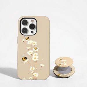 May include: A beige phone case and matching pop-up grip featuring a floral design with white daisies and cartoon bees. The phone case has a black camera lens and black side buttons. The pop-up grip is round and has a similar design.