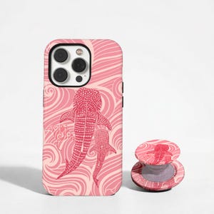 May include: A pink and light pink smartphone case and matching circular phone grip featuring a pattern of whale sharks and manta rays swimming amidst swirling wave lines. The case has a cutout for a triple-lens camera system on the back.
