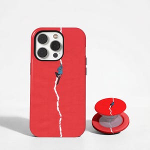 May include: A bright red smartphone case and a matching circular phone grip. Both items feature a vertical white jagged line resembling a paper tear, with a small figure in a blue jacket and black pants climbing along the edge of the white line.