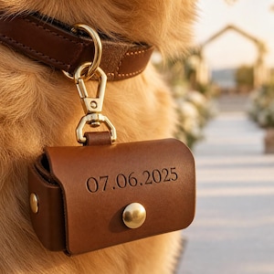 May include: A brown leather dog waste bag dispenser with a gold-colored clasp and a matching brown leather collar. The dispenser is engraved with the date "07.06.2025" and has a gold-colored button closure. The dog's fur is a light brown color.