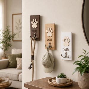 Op de afbeelding: Drie houten wandhaken met pootafdrukontwerpen en dierennamen: "Buddy", "Max" en "Bella". De haken zijn bruin, beige en wit, met metalen haken eronder. Een hondenriem en een pet hangen aan de haken. Een kleine vetplant staat op een tafel.