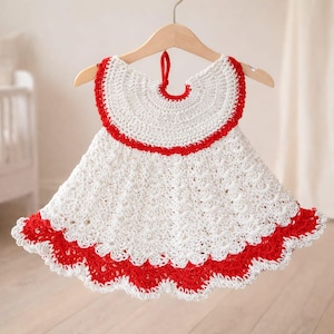 Pode incluir: Vestido branco de crochê com detalhes em vermelho, pendurado em um cabide de madeira. O vestido apresenta uma bainha com babados e um decote com acabamento em vermelho. O vestido é projetado para um bebê ou criança pequena.