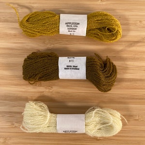 May include: Three skeins of wool yarn in different colors: mustard yellow, dark brown, and cream. The yarn is labeled "Appleton Bros. Ltd. London" and "100% Wool Made in England".