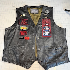 May include: A black leather vest with a V-neck, snap button closures, and an olive green lining. The front features various patches and pins, including Rolling Thunder, Vietnam Veteran, and U.S. Marine Corps insignia, with a Wilsons Leather label.