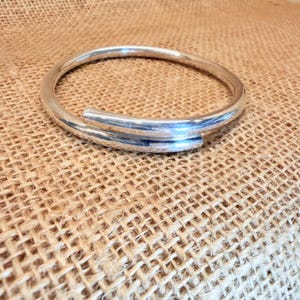 May include: A polished silver-tone bangle bracelet featuring a bypass design where the two ends overlap slightly. The bracelet has a smooth, rounded profile and a reflective metallic finish that catches the light, creating a clean and simple circular form.