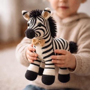 May include: A handmade crochet zebra stuffed animal with black and white stripes, a black mane, and a tan bow tie. The plush toy has black hooves and is being held by a person. The zebra is a soft, cuddly toy.
