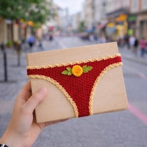 May include: A wooden box wrapped with a red crocheted band designed to resemble underwear. The band is trimmed with a light yellow scalloped edge and features a yellow flower with green leaves. The box is held in a person's hand.