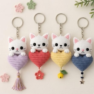 Puede incluir: Cuatro llaveros de gato de ganchillo, cada uno con un gato blanco sosteniendo un corazón. Los corazones son morados, rojos, amarillos y azules. Cada llavero tiene un adorno diferente, incluyendo una borla, una flor, una campana y una estrella.