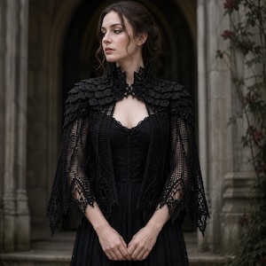 May include: A black crochet bolero featuring a high collar and intricate scale-like patterns on the shoulders, layered over a black dress. The garment has long, flared sleeves with an open-work mesh design and pointed, scalloped edges at the cuffs.