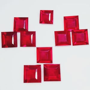 Może przedstawiać: Dziewięć kwadratowych, żywych czerwonych kamieni szlachetnych ułożonych na białej powierzchni. Kamienie mają wypolerowaną, odblaskową powierzchnię i są równomiernie rozmieszczone. Kamienie szlachetne mają głęboki, bogaty czerwony kolor.