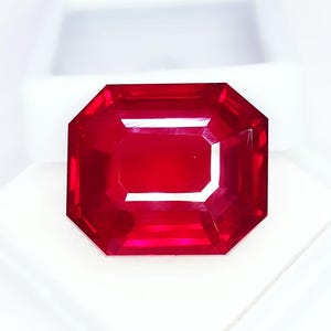 Puede incluir: Una gema roja intensa, tallada en forma rectangular. La gema tiene una superficie brillante y reflectante, sobre un fondo blanco. El tallado de la gema crea múltiples facetas, realzando su brillo. Probablemente un rubí.