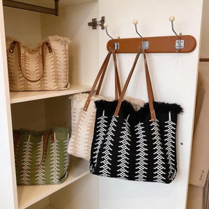 May include: Woven tote bags in beige, black, and green with brown leather handles. The bags have a geometric pattern and fringe trim. One bag is on a shelf, and two are hanging on a brown wooden rack with metal hooks.