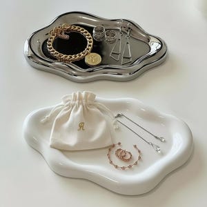 Puede incluir: Dos bandejas decorativas, una plateada y otra blanca, exhiben joyas. La bandeja plateada presenta una pulsera dorada, anillos y pendientes. La bandeja blanca muestra una pequeña bolsa con una "A" y más joyas.