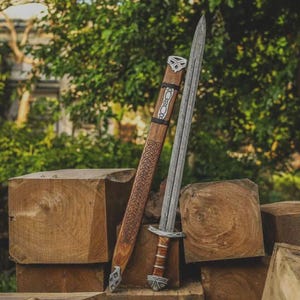 May include: A double-edged steel sword with a leather-wrapped hilt and metal pommel, accompanied by a wooden scabbard featuring intricate carved Celtic knotwork patterns and silver-toned metal accents along the length of the sheath.