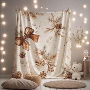 May include: A cream-coloured throw blanket featuring a repeating pattern of brown and tan bows, pinecones, and evergreen sprigs. The design includes subtle speckles across the fabric, creating a festive, nature-inspired aesthetic for the home.