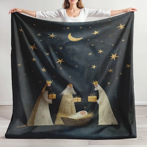 Pode incluir: Um cobertor azul escuro com um design celestial de estrelas douradas e uma lua crescente. O cobertor retrata três figuras com presentes, um bebê em uma manjedoura e uma cena noturna estrelada. O cobertor é segurado por uma pessoa.