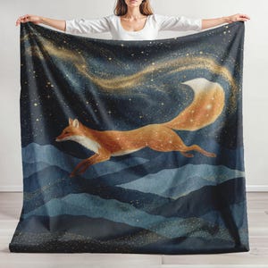 May include: A large rectangular throw blanket featuring a printed design of a fox leaping across a dark blue night sky filled with golden stars and constellations, set above a landscape of layered blue mountains with golden stardust accents.