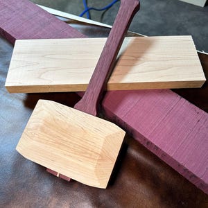 May include: A wooden mallet with a purple wood handle. The mallet is made of light wood and has a square head. The handle is made of a darker wood with a purple hue. The mallet is sitting on a brown leather surface.