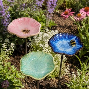Può includere: Tre picchetti da giardino in ceramica a forma di fiore nei colori rosa, blu e verde, montati su una sottile asta di metallo. I piatti rosa e blu presentano una smaltatura lucida e testurizzata, con un'ape che riposa in entrambi.