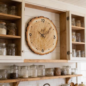 May include: A circular wooden clock with a natural wood grain. The clock face has black numerals at 12, 3, 6, and 9 o'clock, with dots for other hours. The clock hands are dark brown. The clock is mounted on a white wall.