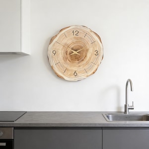 May include: A round wooden clock with a natural wood grain pattern. The clock face features black numerals and hands, with a light brown and beige color scheme. The clock is mounted on a white wall.