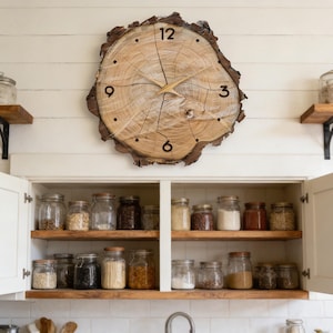 May include: A large, round wooden clock with a natural edge is mounted on a white wall. The clock face features black numerals and gold-colored hands. Below, a white cabinet with open doors reveals shelves filled with glass jars.