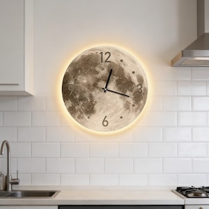 May include: A round wall clock with a moon design. The clock face features a detailed lunar surface with the numbers 12, 3, 6, and 9. The clock has black hands and a warm, glowing light around the perimeter. It is mounted on a white tiled wall.