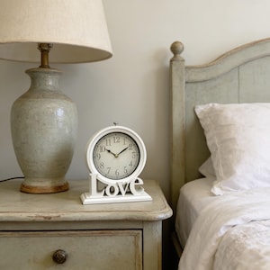 Op de afbeelding: Een witte, ronde klok met zwarte cijfers en wijzers staat op een nachtkastje. De basis van de klok spelt het woord "Love" in witte letters. Een beige lamp en een bed met een wit kussen zijn op de achtergrond.