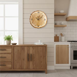 May include: A round wooden wall clock made from a cross-section of a tree trunk. The clock face features black numerals at the 12, 3, 6, and 9 o'clock positions, with thin black hands. The clock is mounted on a light beige wall.