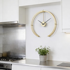 May include: A gold-colored, open-faced wall clock with black hands hangs on a white wall in a kitchen. The clock has a minimalist design with a circular shape. A small potted plant sits on the countertop below the clock.