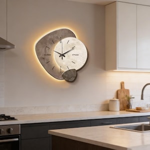 Puede incluir: Un reloj de pared moderno con un diseño único. El reloj presenta una esfera circular con manecillas negras y una apariencia texturizada similar a la piedra. El reloj está rodeado por una forma gris abstracta con luz cálida.