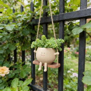 May include: A whimsical hanging planter featuring a smiling face design. The beige ceramic pot holds a vibrant green succulent and is suspended by rope. The planter has arms and legs, and sits on a wooden swing.