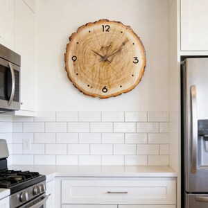 May include: A large, round wall clock made from a cross-section of a tree trunk. The clock face features natural wood grain, black numerals, and black clock hands. The clock is mounted on a white wall in a kitchen setting.