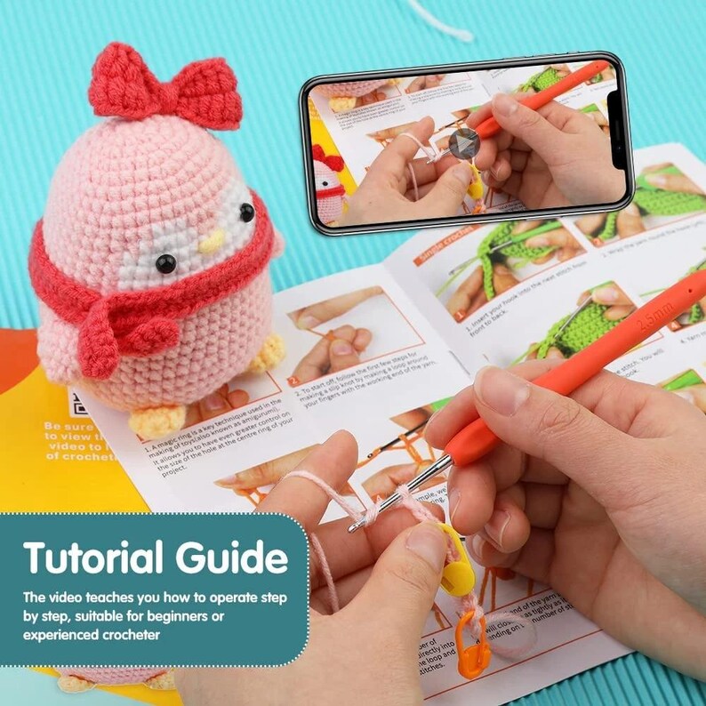 May include: A pink and red crocheted penguin with a bow and scarf sits next to a tutorial guide. The guide shows step-by-step instructions for crocheting, with an orange crochet hook and yarn visible. The text on the guide says "Tutorial Guide".