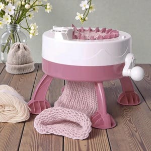 May include: A white and pink knitting machine with a light beige knitted hat and a roll of yarn. The machine has a white handle and is on a wooden surface. A small vase of white flowers is in the background.