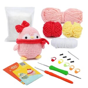May include: A crochet kit featuring a pink penguin with a red bow and scarf. The kit includes yarn in pink, red, yellow, and white, along with crochet hooks, stitch markers, stuffing, and instructions. A DIY craft project.