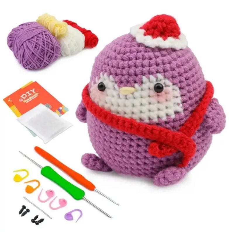 May include: A DIY crochet kit featuring a purple penguin with a red scarf and Santa hat. The kit includes yarn, crochet hooks, stitch markers, and a small instruction booklet. The penguin is approximately 5 inches tall.