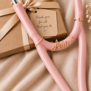 Op de afbeelding: Een roze, Y-vormig decoratief object met gouden draadaccenten en de naam "Aria". Een geschenkdoos met een strik en een label met de tekst "Thank you for all that you do" staat op de achtergrond. Het object heeft een gladde, glanzende afwerking.