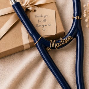以下が含まれることがあります： 「Madison」という名前を含む、ゴールドのアクセントが付いたネイビーブルーの聴診器。背景には、ベージュのリボンが付いたギフトボックスと、「Thank you for all that you do」と書かれたタグがあります。