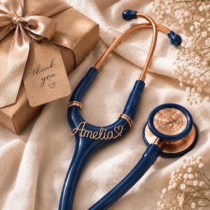 Puede incluir: Un estetoscopio azul marino con detalles en metal oro rosa, que presenta el nombre Amelia en letra cursiva y un pequeño símbolo de corazón unido al tubo. Una caja de regalo con una etiqueta color canela que dice thank you con un corazón.
